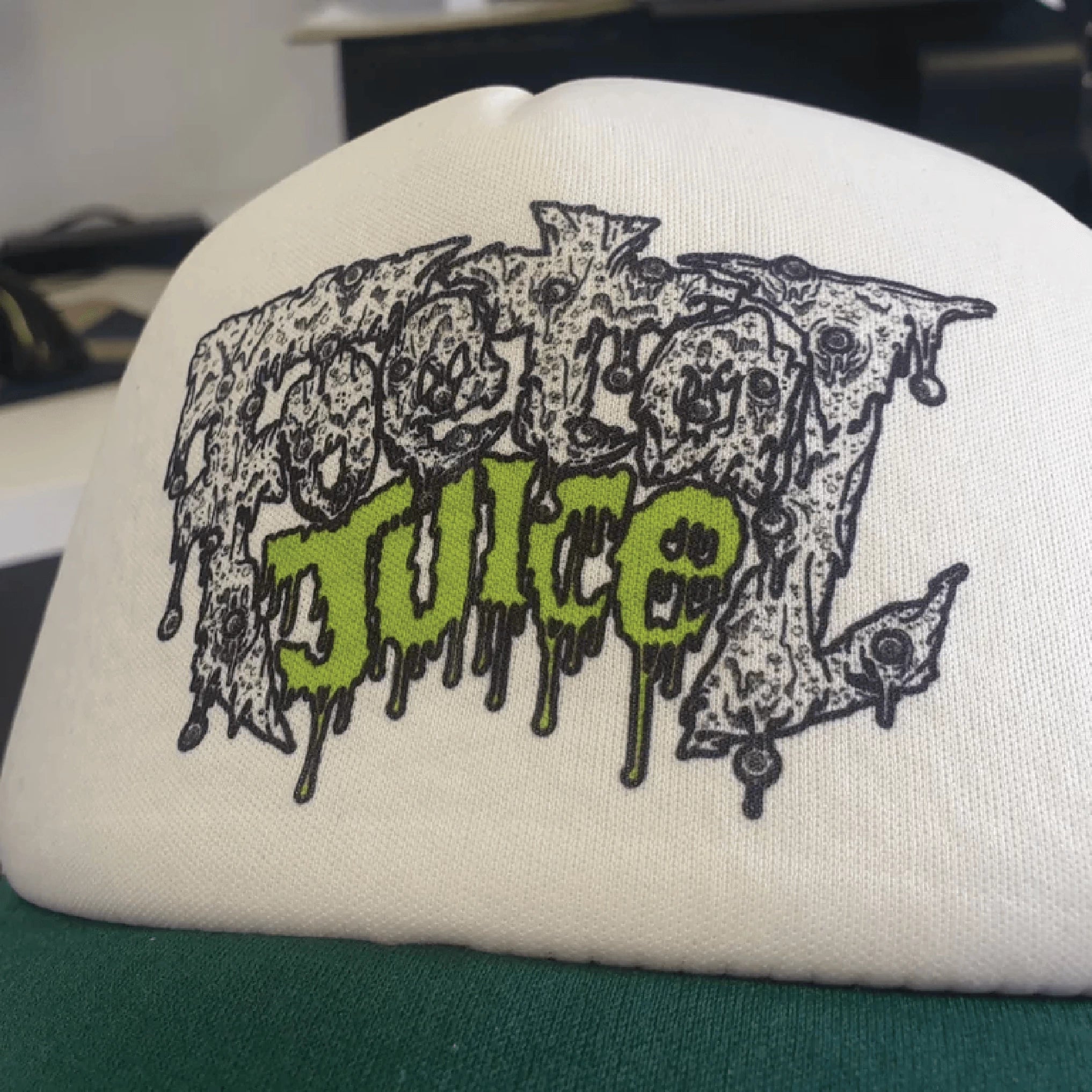 Green Foetal Juice trucker cap with a close up of the logo print on the front.
