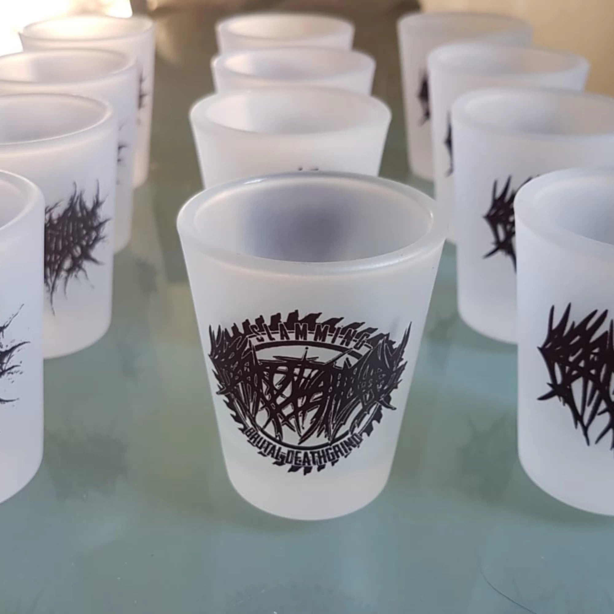 Frosted shot glasses with a black band logo on a reflective surface