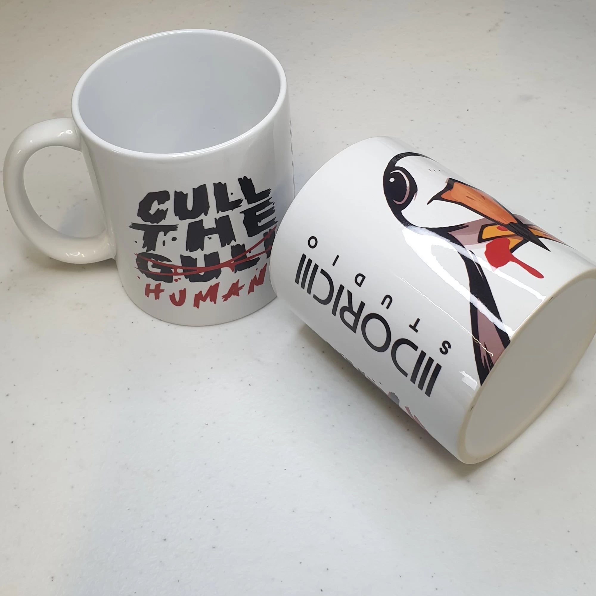 Two mugs on a white surface, one with a cull the humans design and the other with a bird design.